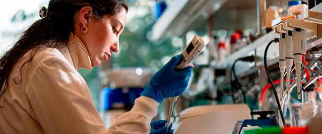 A woman in a white lab coat wearing blue lab gloves sits in a lab using equipment