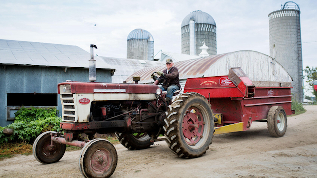 Farmer on tractor pulling some machinery with shop and grain silos in the background.